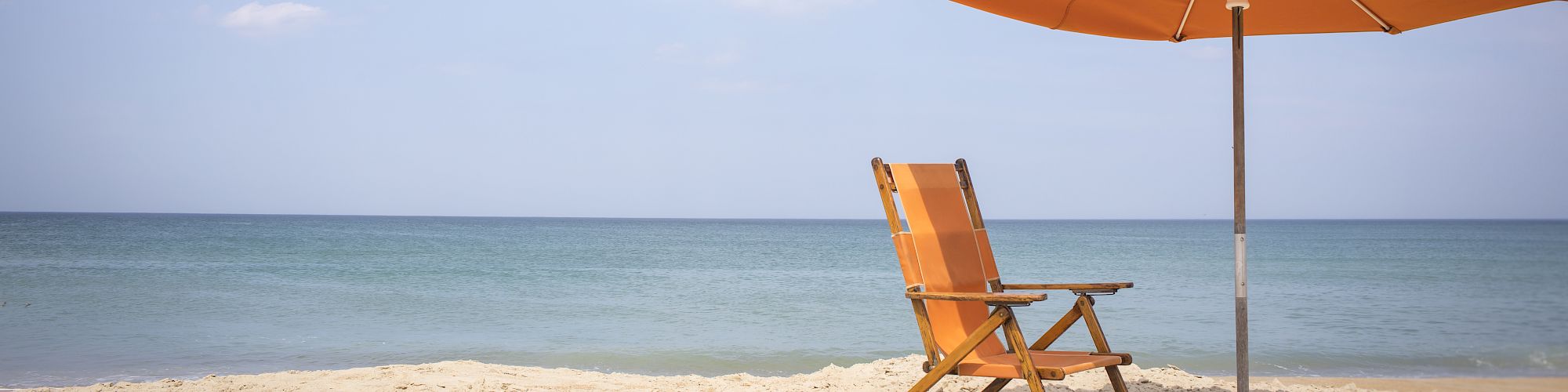 An orange beach chair and umbrella are set up on a sandy beach overlooking a calm, blue ocean under a partly cloudy sky.