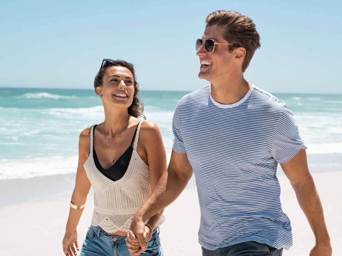 A couple is walking hand in hand on a sandy beach, smiling and enjoying the sunny day by the ocean.