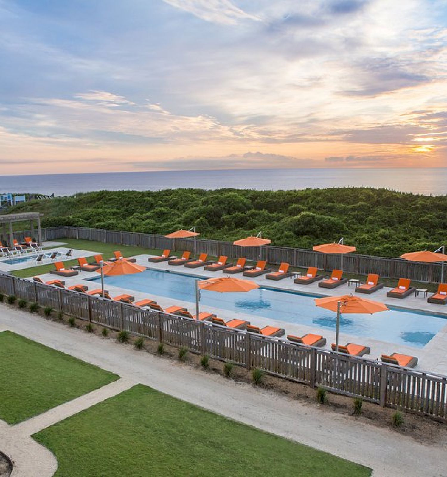 A pool area with orange parasols and lounge chairs, surrounded by greenery and a rustic fence, overlooking a serene ocean sunset.