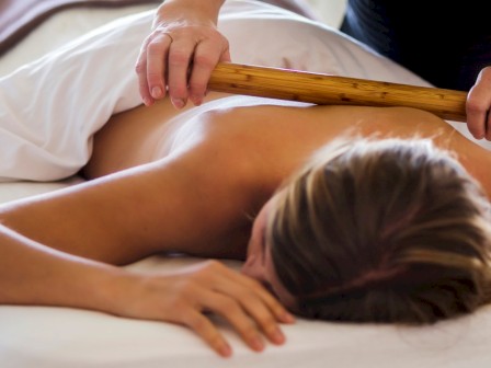 A person is lying face down receiving a bamboo massage on their back, covered partially with a white sheet.