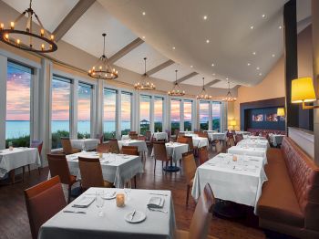 A spacious restaurant with large windows, white tablecloths, and ambient lighting offers a scenic view of the sunset over the ocean.