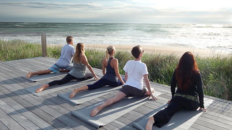 Five people are doing yoga on mats on a wooden deck near the beach, facing the ocean during a serene, sunny day.