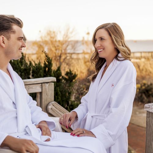Two people in white robes are smiling and conversing outside on a wooden deck with a scenic background.