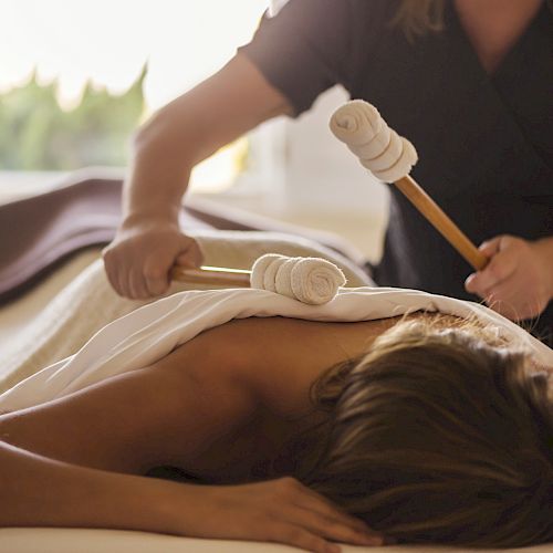 A person receiving a massage with the therapist using wooden tools wrapped in cloth on their back.