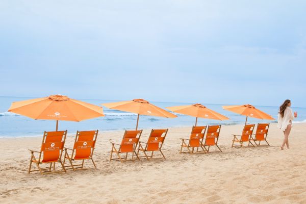 An image of a beach with a row of empty orange chairs and umbrellas. A person walks nearby, with the ocean waves visible in the background.
