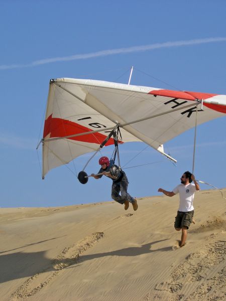 A person is hang gliding, taking off from a sandy area, while another person runs alongside holding the glider's frame.