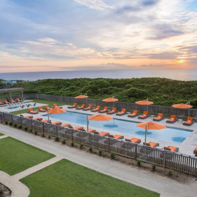 A serene pool area with orange umbrellas and lounge chairs, surrounded by a wooden fence, sits adjacent to a green landscape with an ocean view.