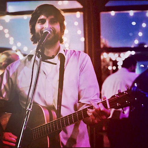 A man is playing an acoustic guitar and singing into a microphone at a cozy venue with string lights and people in the background.