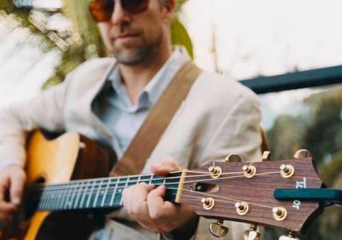 A person wearing sunglasses and a light-colored jacket is playing an acoustic guitar outdoors, with a blurred background of greenery.