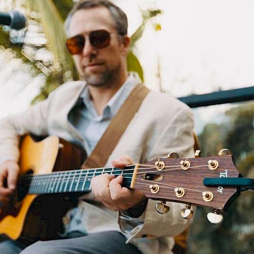 A person wearing sunglasses and a light-colored jacket is playing an acoustic guitar outdoors, with a blurred background of greenery.