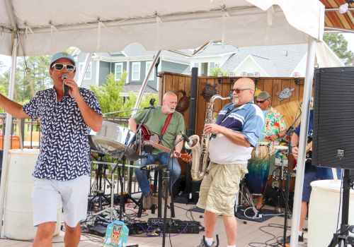 A band is performing on a small outdoor stage with a singer, guitarist, saxophonist, and drummer under a tent.