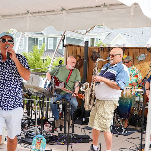 A band is performing on a small outdoor stage with a singer, guitarist, saxophonist, and drummer under a tent.