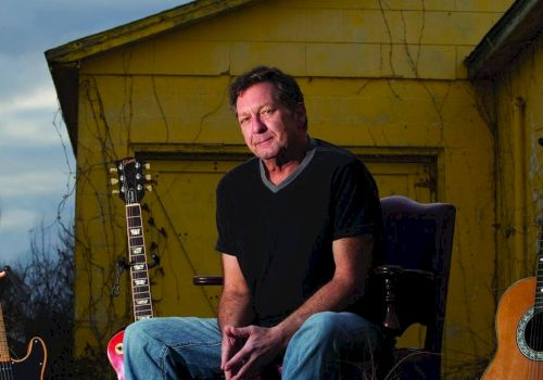 A man sits in front of a yellow house surrounded by guitars on the ground, with a cloudy sky in the background.