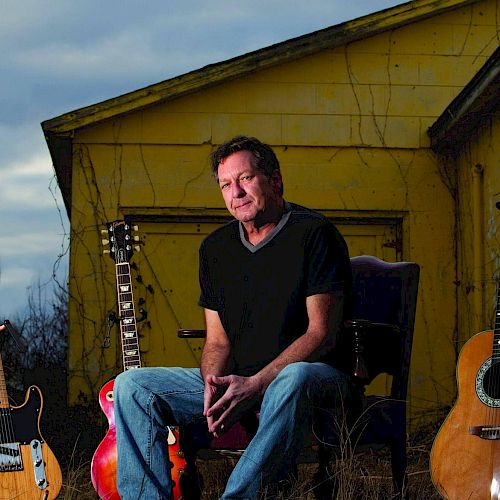 A man sits in front of a yellow house surrounded by guitars on the ground, with a cloudy sky in the background.