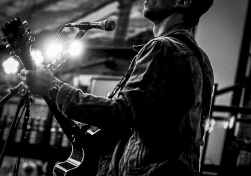 A black-and-white image shows a person playing a guitar, singing into a microphone, and wearing a jacket. There are visible lights in the background.