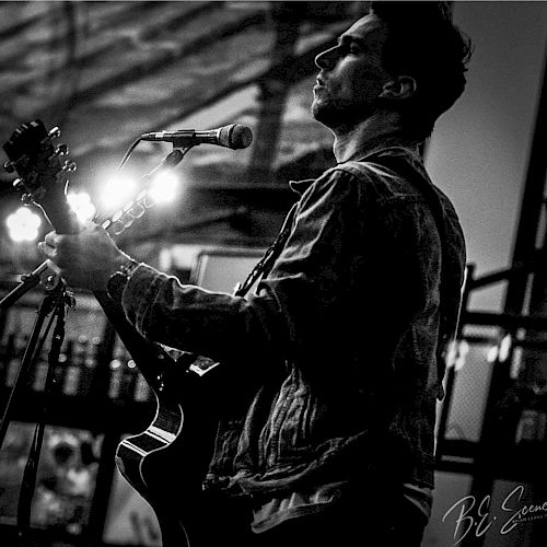 A black-and-white image shows a person playing a guitar, singing into a microphone, and wearing a jacket. There are visible lights in the background.