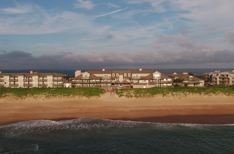 A beachfront resort with multiple buildings, situated along a sandy beach with waves washing ashore and a partly cloudy sky above.