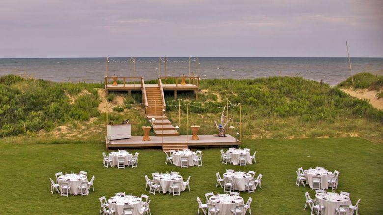 Outdoor event setup with round tables and chairs on green grass, a raised wooden platform with stairs, and the ocean in the background.