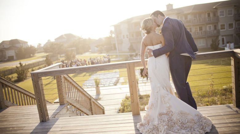 A couple in wedding attire embraces on a wooden deck overlooking a lawn and buildings in the background.
