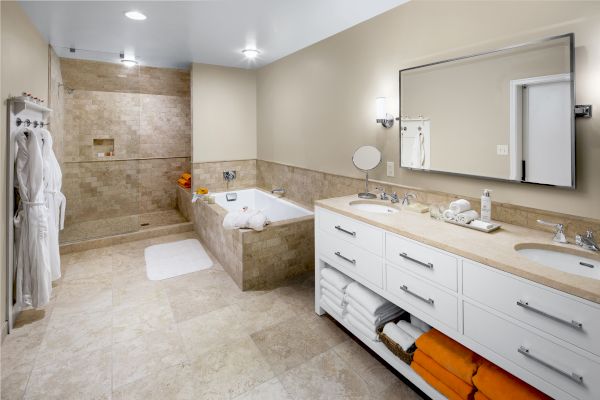 A modern bathroom with a large walk-in shower, bathtub, double sink vanity, mirror, towels, robes, and tiled floor in neutral tones.