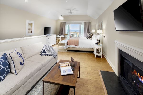 A cozy room features a bed, sofa, coffee table, fireplace, two TVs, and a balcony with an ocean view, adorned in neutral tones and nautical accents.