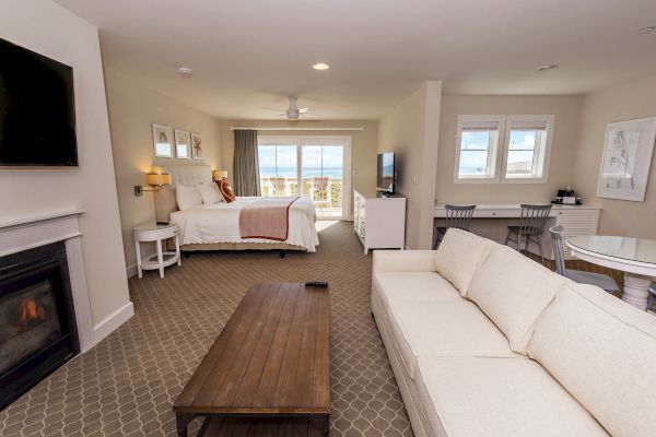 A spacious hotel room with a bed, couch, coffee table, fireplace, TV, and a balcony view of the ocean. Light and airy interior design.