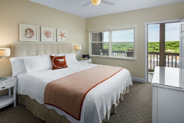 A bright, cozy bedroom features a large bed with a beige headboard, ocean-themed decor, and a view of greenery and water through the window and balcony door.