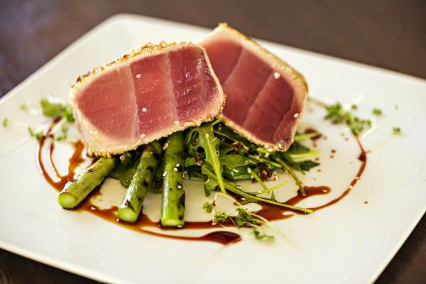 The image shows a gourmet dish featuring seared tuna slices atop a bed of asparagus and greens, served with a drizzle of balsamic reduction.