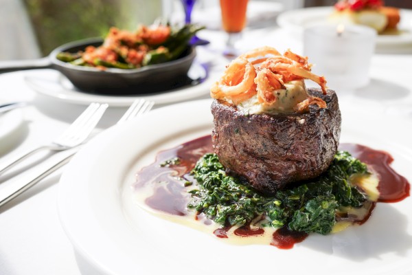 A plated steak dish with sautéed greens, garnished with crispy onions. A side dish is in the background on a table set with utensils.