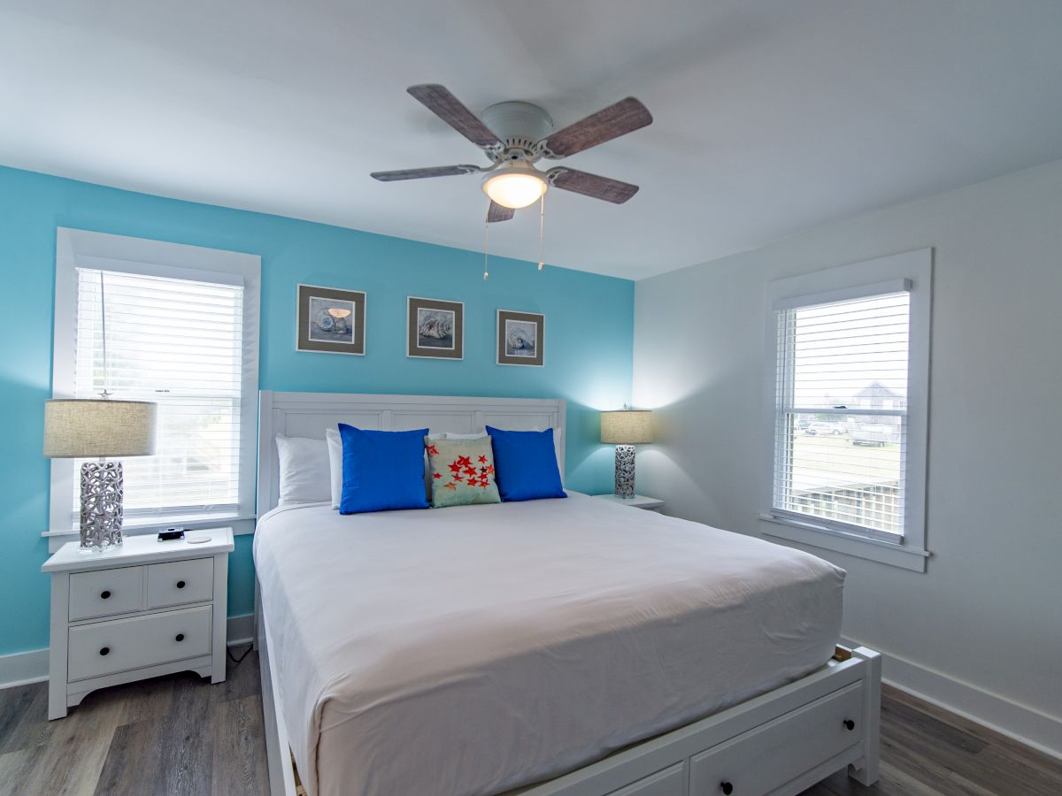 A bright bedroom with blue accent wall, large bed with white linens, blue pillows, two nightstands, lamps, three pictures, and a ceiling fan.