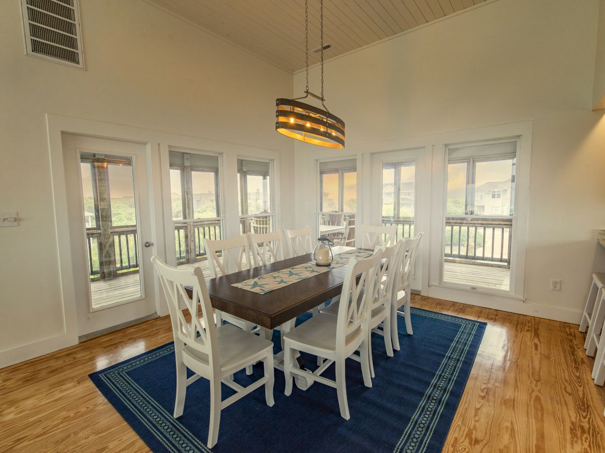 A dining room with a rectangular table, white chairs, a hanging light fixture, and large windows, featuring a blue rug and wooden flooring.