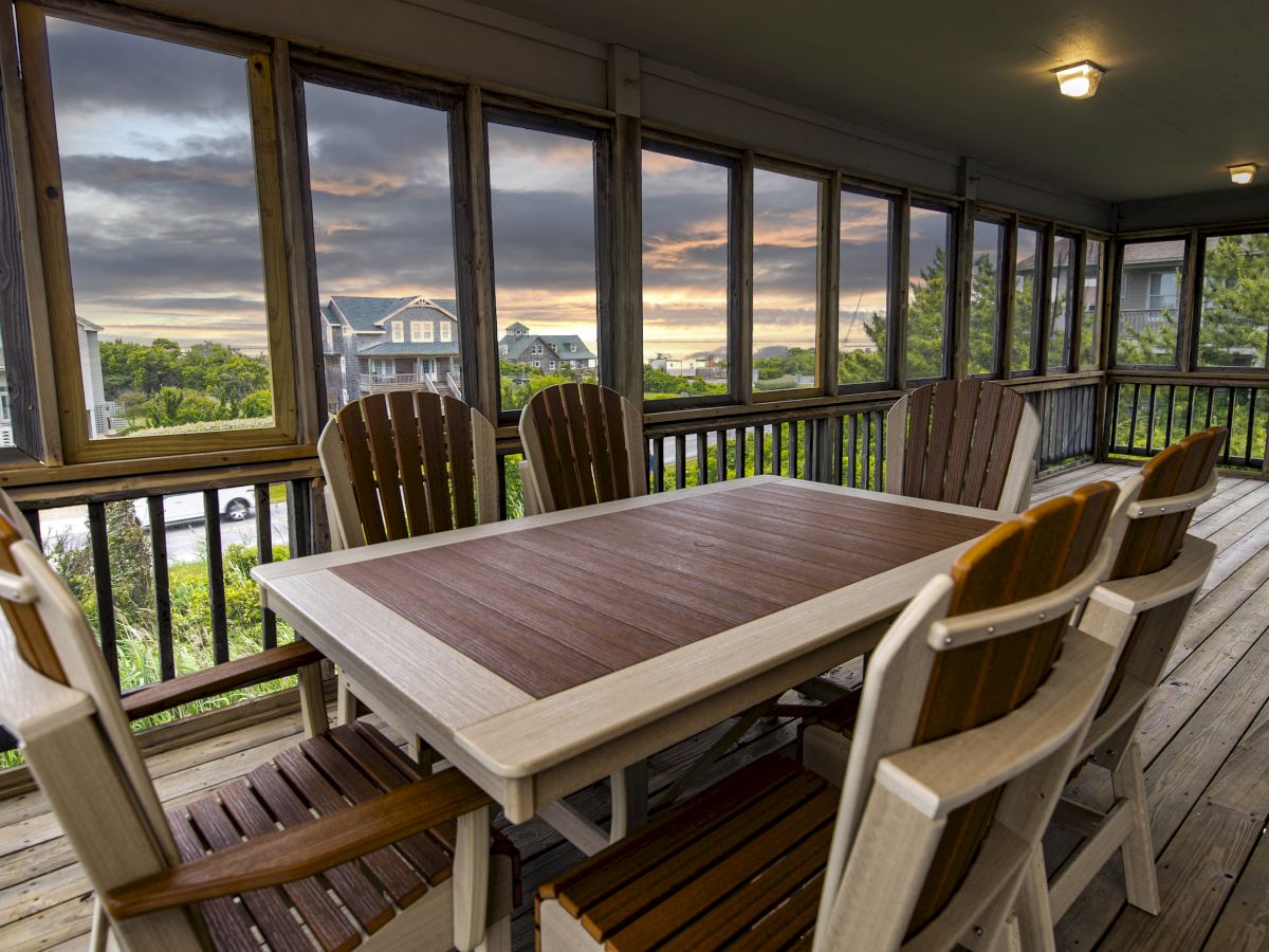 A screened porch with a wooden dining table and six chairs, overlooking a scenic view with houses and greenery. The sun sets in the background.