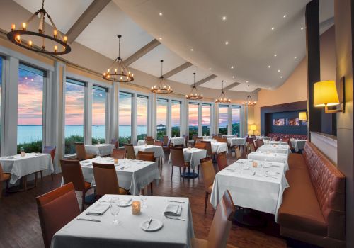 An elegant restaurant with large windows overlooking a scenic sunset, set tables, cushioned seating, and chandelier lighting creates a cozy atmosphere.