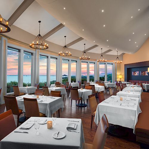 An elegant restaurant with large windows overlooking a scenic sunset, set tables, cushioned seating, and chandelier lighting creates a cozy atmosphere.