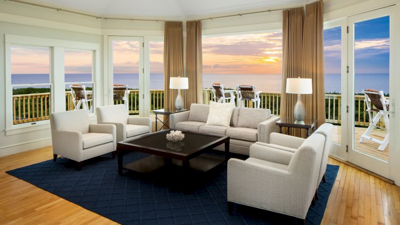 A modern living room with beige sofas, a dark coffee table on a blue rug, and windows showing a scenic ocean view at sunset.