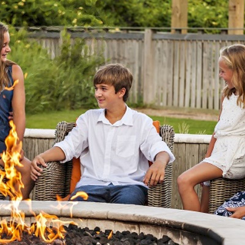 A family of five is sitting around a fire pit in their yard, smiling and enjoying each other's company on a sunny day in a grassy backyard.