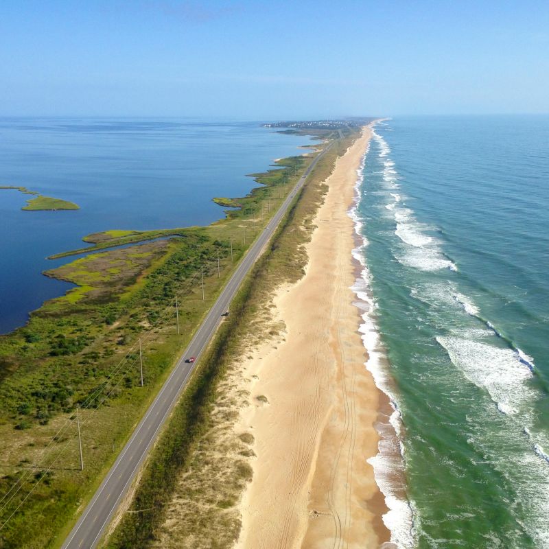 An aerial view of a coastal road flanked by a sandy beach and ocean on one side, with a body of water and green land on the other side.