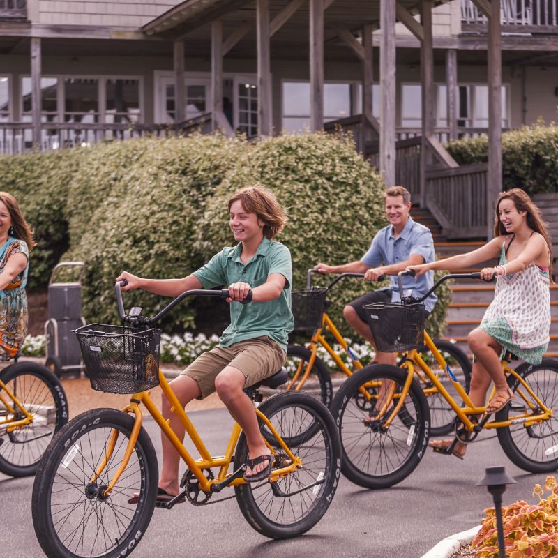 Four people are riding yellow bicycles together on a paved path, with a wooden building and greenery in the background.