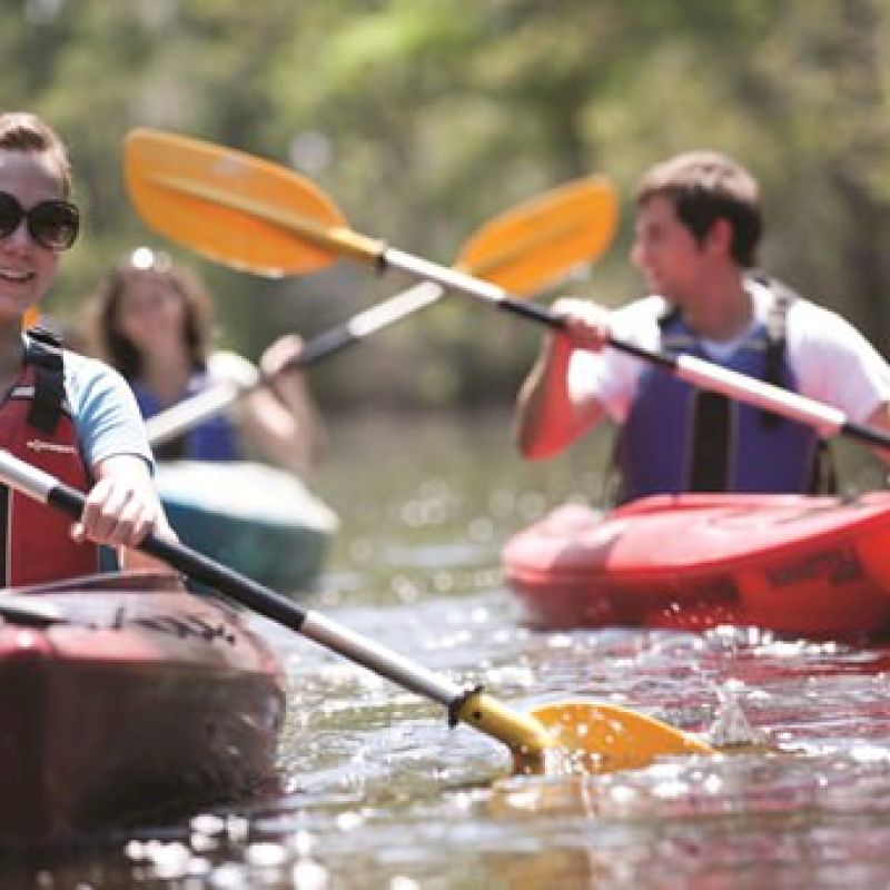 Three people are kayaking on a calm body of water, wearing life jackets and enjoying a sunny day outdoors.