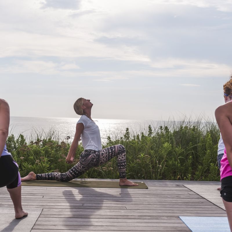 A group of people practice yoga outside on a wooden deck near the beach with serene scenery and ocean in the background.