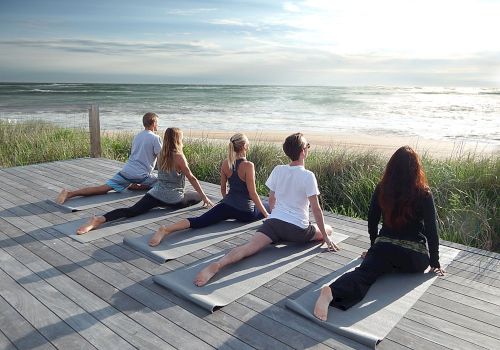 A group of five people is practicing yoga on mats overlooking the ocean on a wooden deck.
