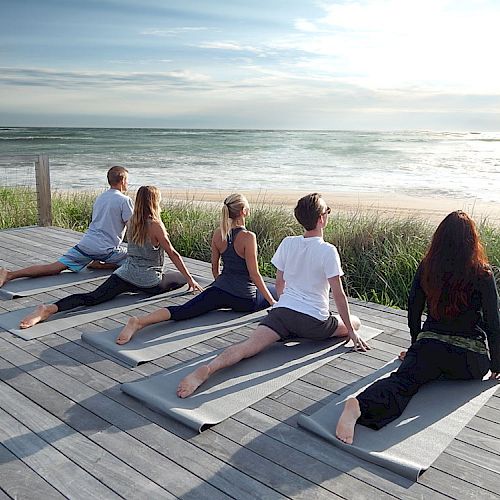 A group of five people is practicing yoga on mats overlooking the ocean on a wooden deck.