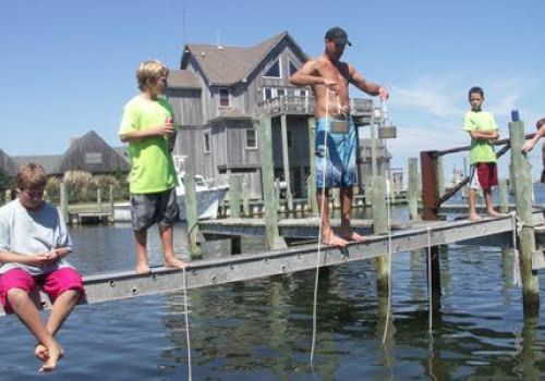 A group of people are standing and sitting on a dock, possibly fishing or crabbing, with houses and water in the background.