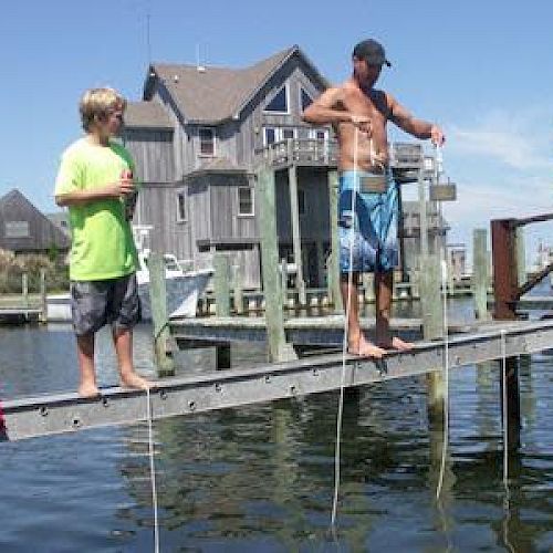 A group of people are standing and sitting on a dock, possibly fishing or crabbing, with houses and water in the background.