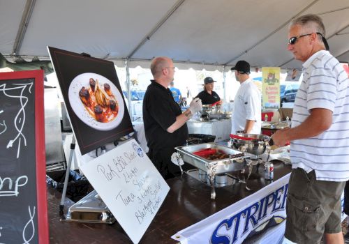 A man at a food stall talking to chefs under a tent with a large food photo display and dish sign, and a blackboard to the side ends the sentence.