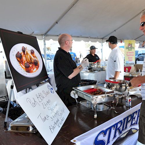 A man at a food stall talking to chefs under a tent with a large food photo display and dish sign, and a blackboard to the side ends the sentence.