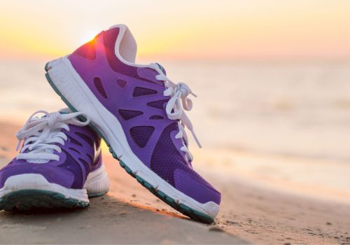 A pair of purple and white athletic shoes sits on a sandy beach with the ocean and sunset in the background.