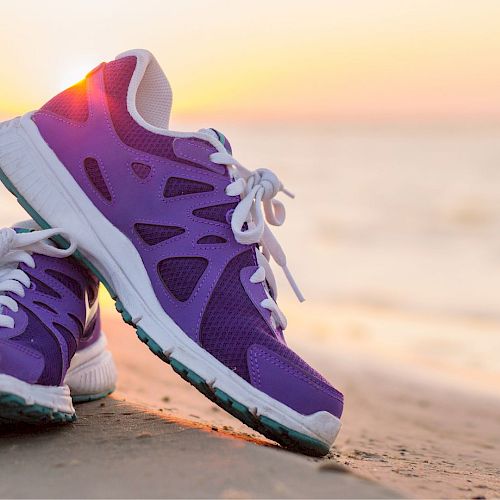 A pair of purple and white athletic shoes sits on a sandy beach with the ocean and sunset in the background.