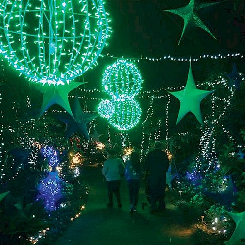 People walking through a beautifully decorated garden with glowing green and blue lights, star-shaped lanterns, and hanging spherical ornaments at night.