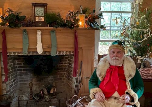 A man dressed as Santa Claus sits in a festive room with a fireplace, stockings, a Christmas tree, and holiday decorations, holding a musical instrument.